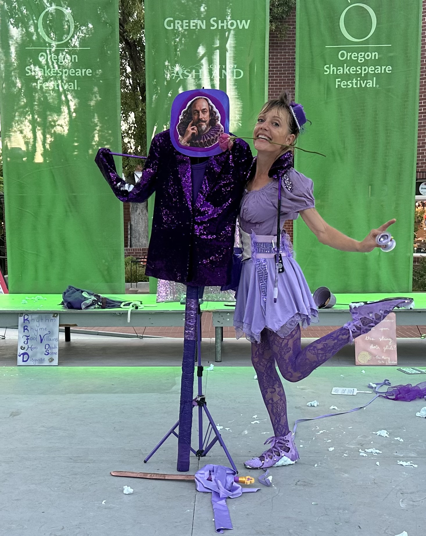 Jenifer Joy holding a rose in her teeth beside the Shakespeare costume stand at the Oregon Shakespeare Festival Green Show