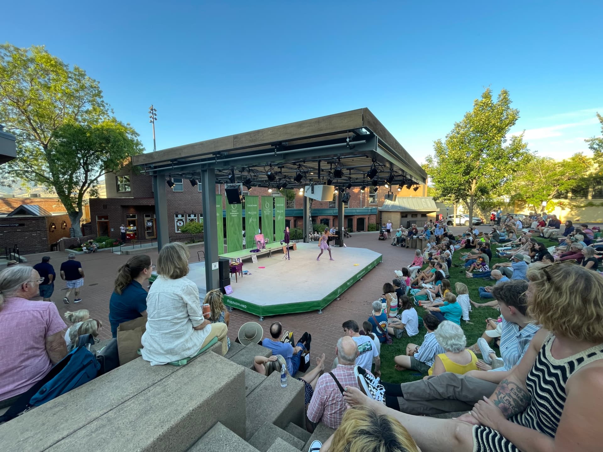 Wide view of the Oregon Shakespeare Festival Green Show stage with a full audience watching The Merry Yo-Yo of Windsor