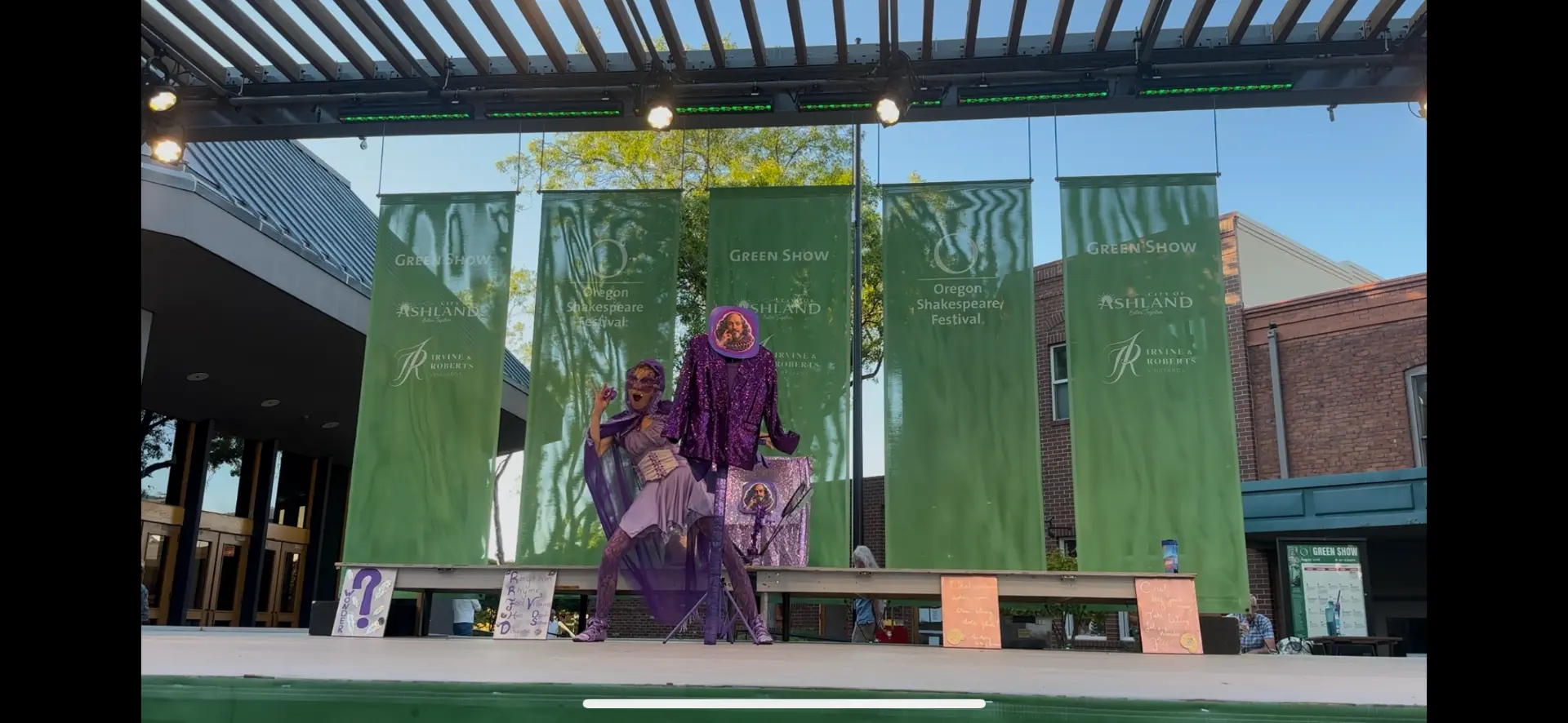 Jenifer Joy posing beside the Shakespeare costume stand on the Green Show stage in Ashland