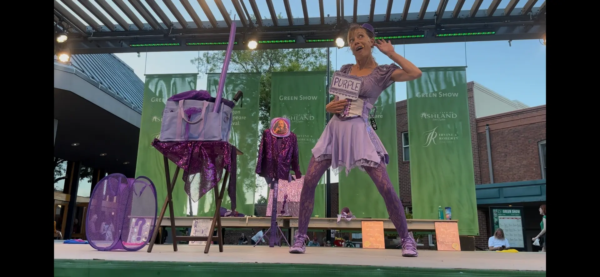 Jenifer Joy holding a hand-lettered PURPLE sign and cupping her ear for the audience